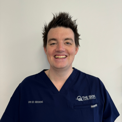 Dr Eddy Siddens smiles at the camera in front of a white background. He has short dark hair and is wearing navy blue scrubs